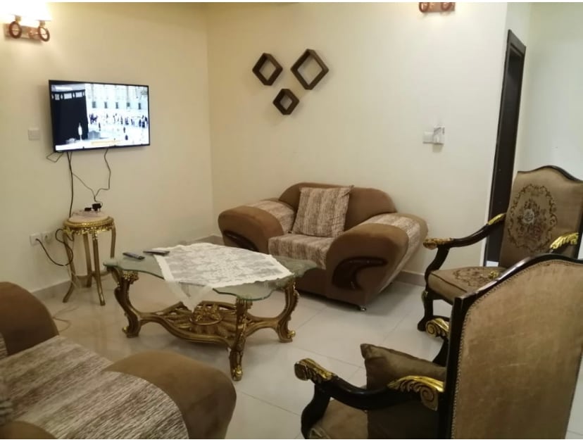 A cozy seating area features a plush brown sofa set with decorative cushions, complemented by a glass coffee table. A wall-mounted television is visible, and a few decorative wall frames add interest. Light-colored walls enhance the inviting atmosphere.