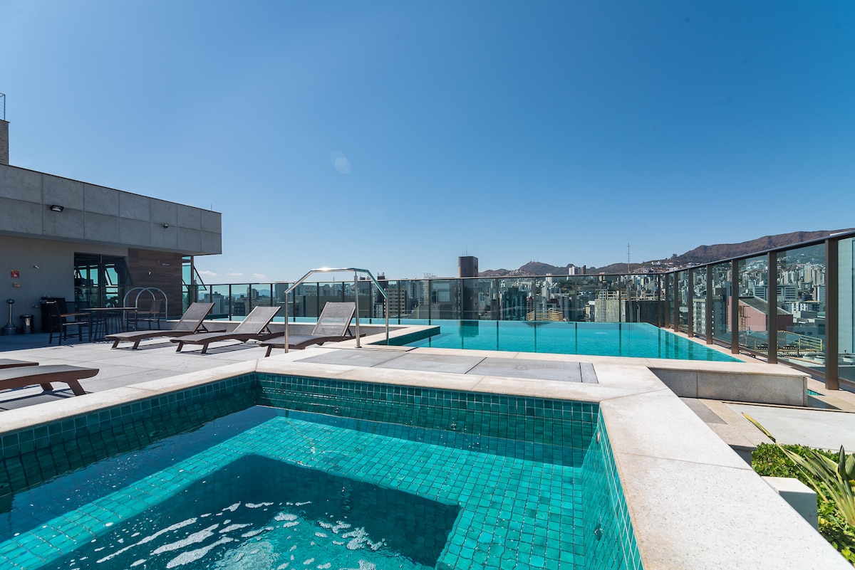 A rooftop area features an infinity pool alongside a jacuzzi. Lounge chairs are positioned around the pool, offering relaxation with panoramic city views. The glass railing allows for unobstructed sightlines, emphasizing the open and airy atmosphere of the space.
