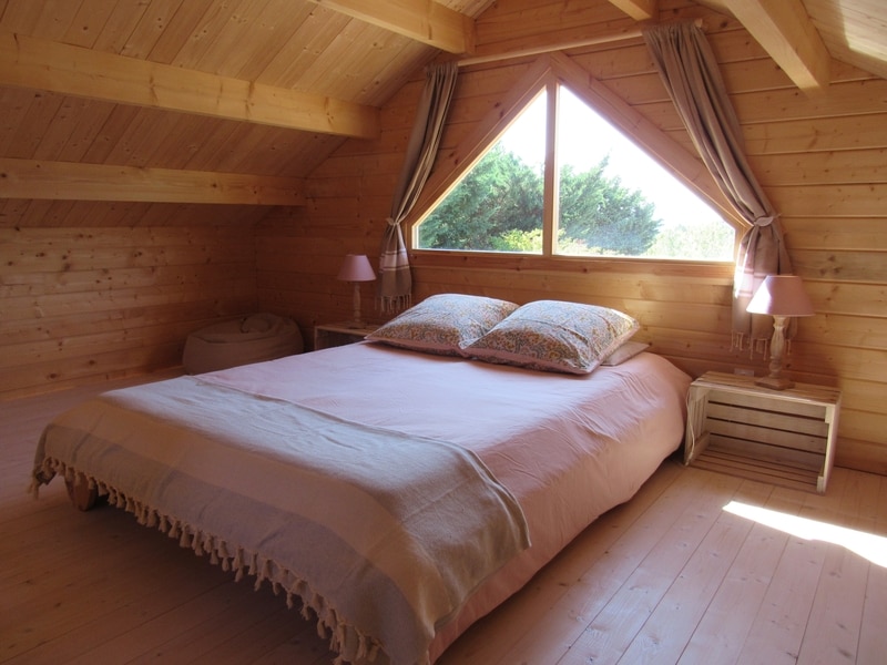 A wooden loft bedroom features a spacious bed adorned with soft bedding and decorative pillows. Natural light filters through a large triangular window, illuminating the warm wooden walls. Two gentle pink lamps are positioned on either side of the bed, contributing to the welcoming ambiance.