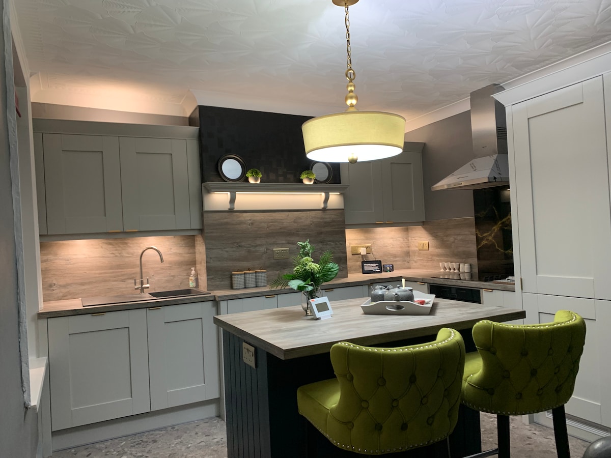 The kitchen features modern cabinetry with a neutral palette, illuminated by soft lighting. A central island with two green upholstered chairs offers a casual dining space, while chic decor accents and stainless steel appliances complete the stylish design.