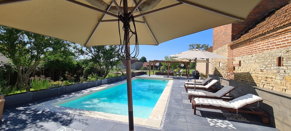 A serene outdoor space features a refreshing swimming pool surrounded by comfortable lounge chairs. Sun umbrellas provide shade, while lush greenery adorns the perimeter, enhancing the relaxing atmosphere. An inviting pool house is visible in the background, contributing to the tranquil setting.