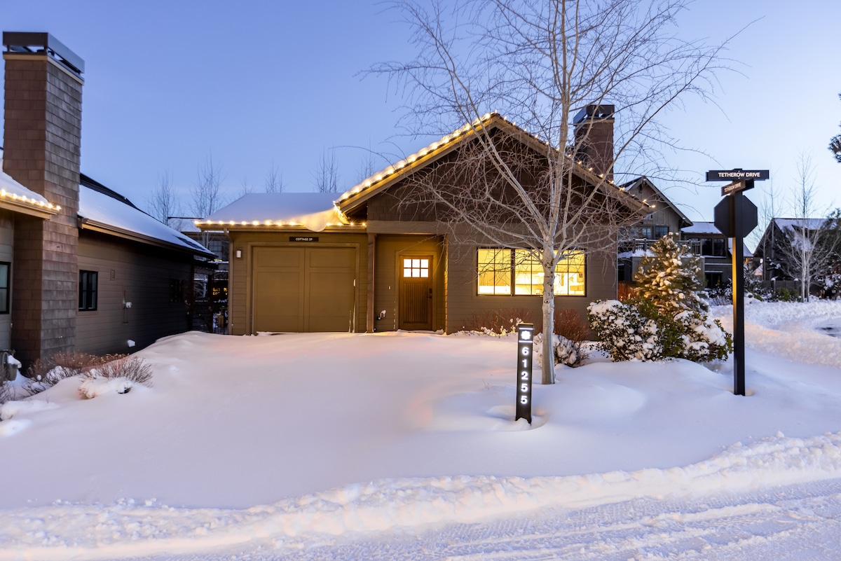 A cozy cottage exterior is covered with fresh snow, accented by soft outdoor lighting. The front features a warm yellow glow from the windows, and a garage door is visible to the left. A signpost stands near the entrance, indicating the street name.