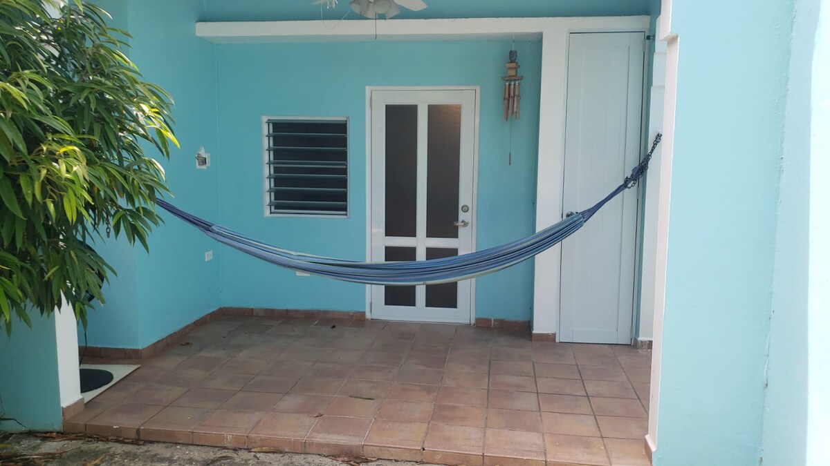 A relaxed outdoor space features a blue and gray hammock suspended between two walls. The inviting area is accented by light blue walls and a tiled floor, with a door and window framed in white providing access. Lush greenery enhances the tranquil atmosphere.