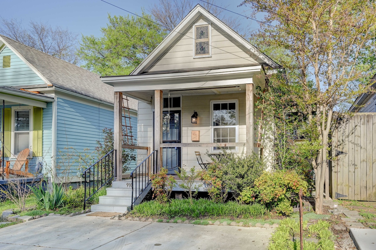 A charming bungalow entrance is showcased, featuring a front porch with a small table and chairs. Lush greenery surrounds the steps, enhancing the inviting façade of the home. Adjacent properties are visible, reflecting a colorful and vibrant neighborhood atmosphere.