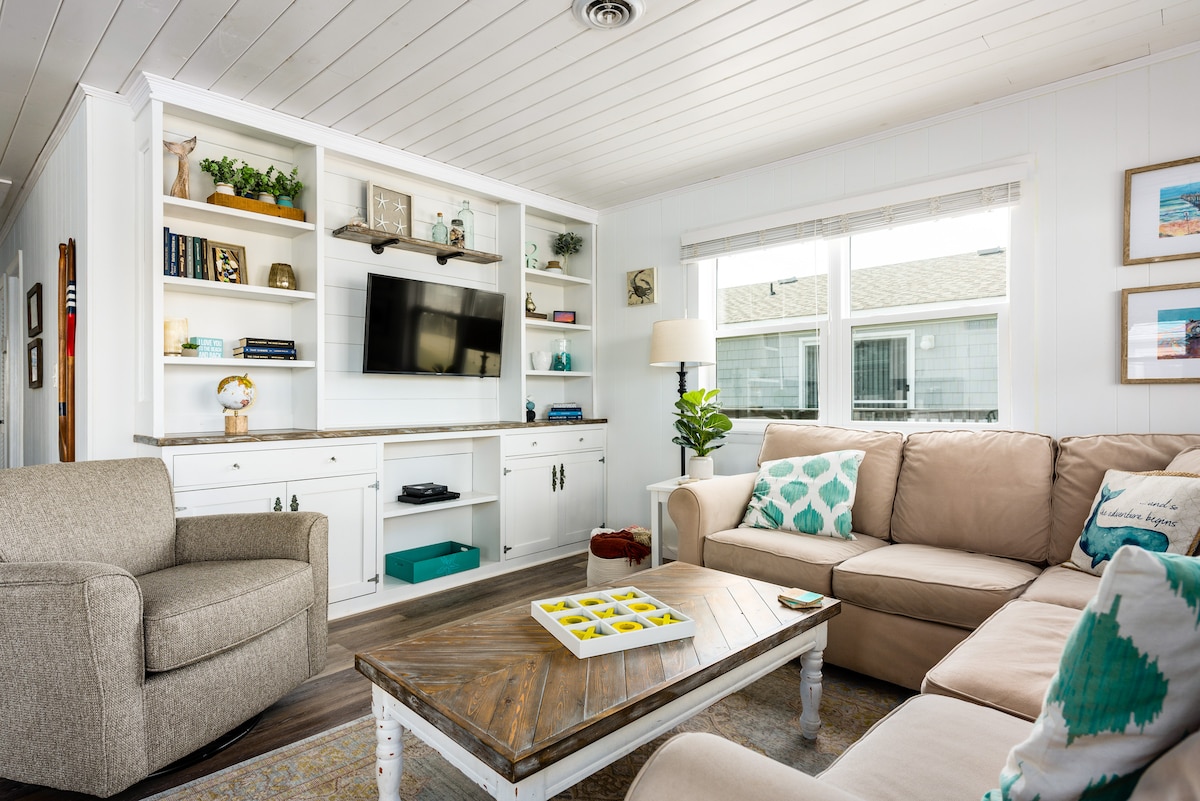 A bright living area is presented with a beige sectional sofa and a gray armchair. A wooden coffee table sits at the center. A white entertainment unit with bookshelves houses a smart TV and decorative items. Natural light filters in through a large window, enhancing the open feel.