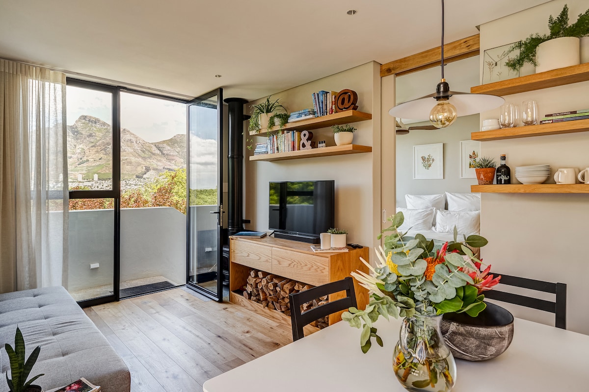 An open-plan living area is shown, featuring a dining table, stylish shelves displaying books and decor, and a modern television setup. Large glass doors create a connection to the outdoors, with views of nearby mountains and a cozy log storage area.