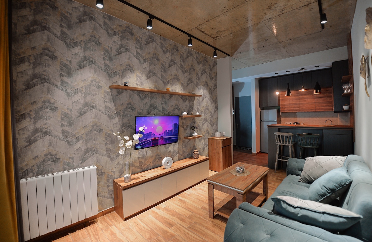 The living area features a modern decor with a patterned wall, wooden shelves, and a mounted television. A comfortable sofa faces the TV, while a coffee table sits in front. The open layout connects to a kitchen area with bar seating, illuminated by pendant lights.