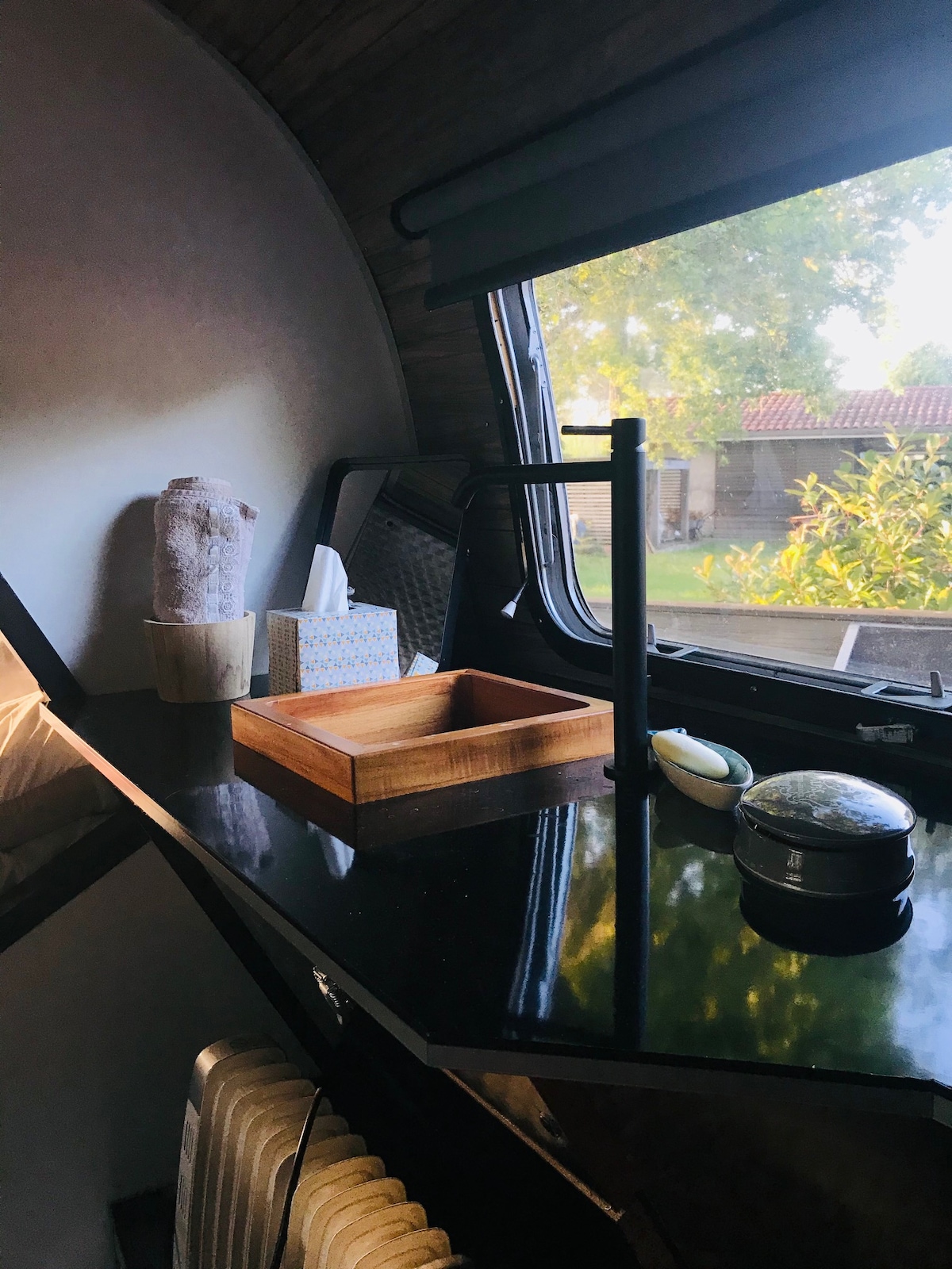 A wooden tray sits atop a dark countertop, accompanied by a glass jar and a soap dish. Nearby, rolled towels are placed above a tissue box. Natural light filters through a window, illuminating the space and highlighting surrounding greenery.