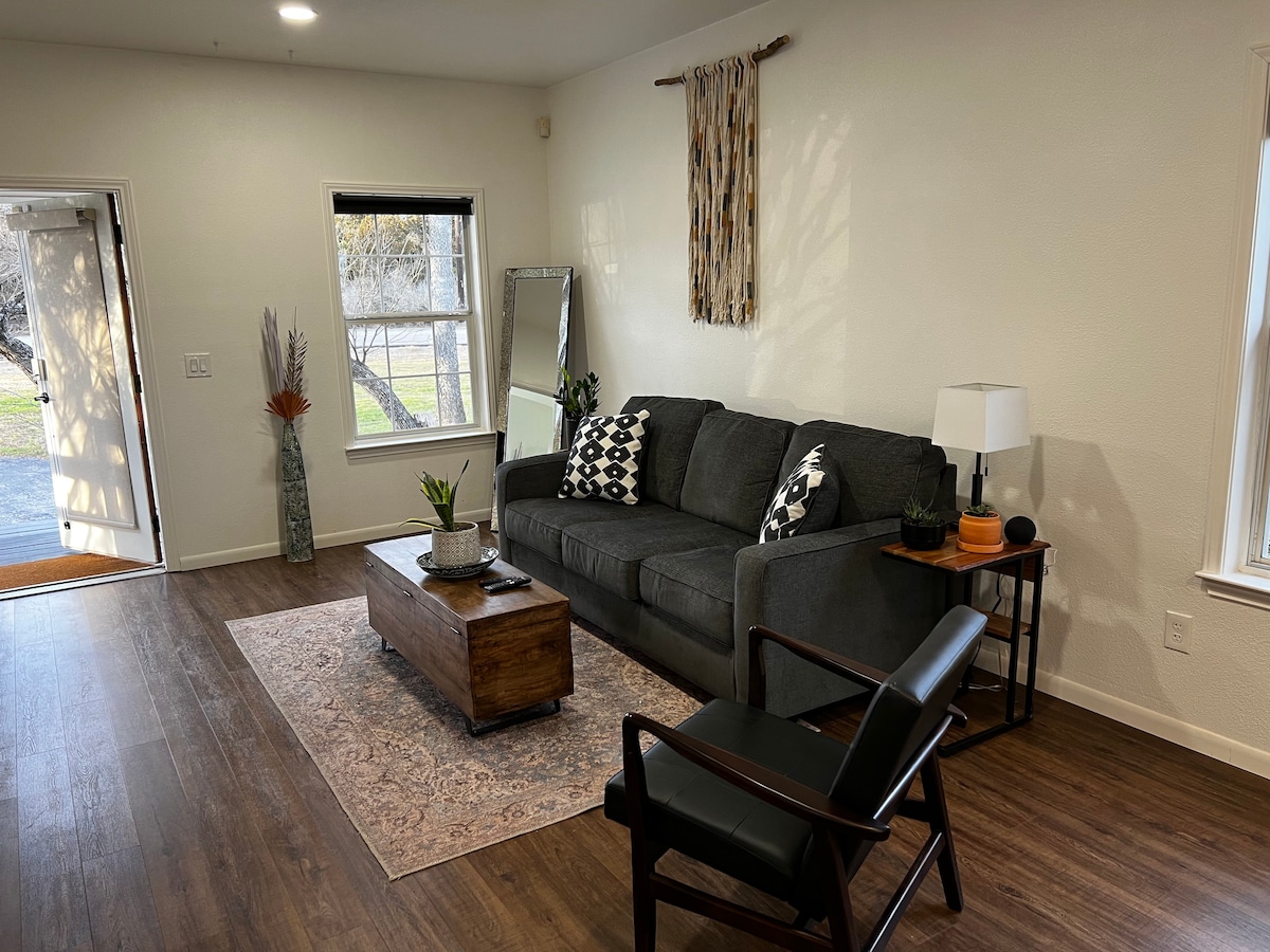 A spacious living area features a comfortable gray sofa adorned with patterned cushions. A wooden coffee table sits in front, accompanied by a dark armchair. Natural light enters from the nearby window, highlighting a potted plant and decorative accents.