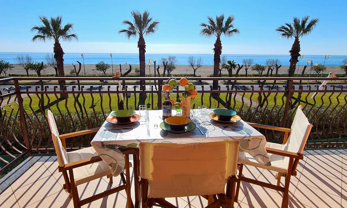 A balcony table is set for a meal, featuring two plates and vibrant flowers as a centerpiece. Palms sway in the background, framing the view of the sea and sandy beach, with a clear blue sky above, offering a serene outdoor dining space.