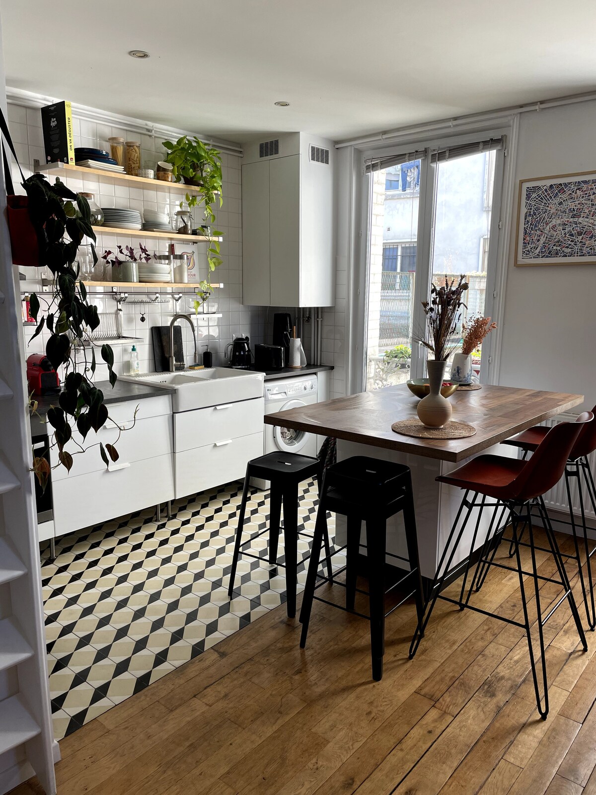 A bright kitchen area is shown, featuring modern white cabinetry and open shelving displaying dishware and plants. A wooden table with four stools invites dining, while a geometric patterned floor adds visual interest. Natural light filters through large windows, illuminating the space.
