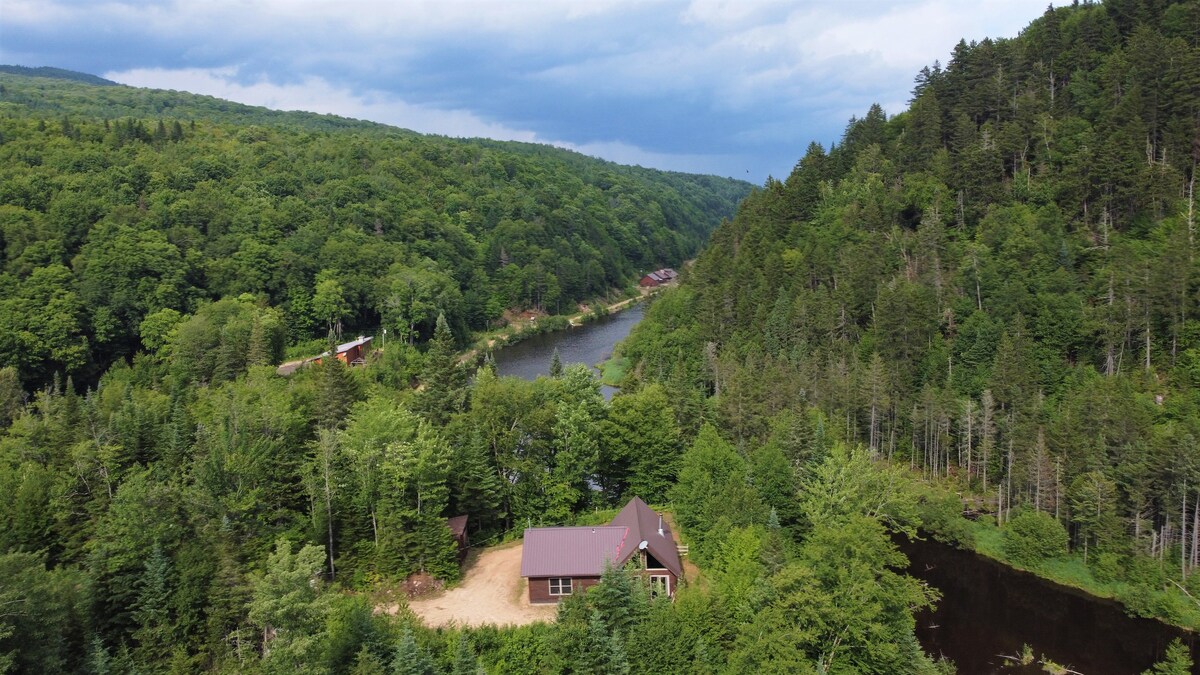 Aerial view of a secluded chalet nestled among lush greenery, alongside a winding river. Surrounding mountains and dense forests create a natural backdrop, showcasing the beauty of the area. The chalet features a brown roof, blending harmoniously with the environment.