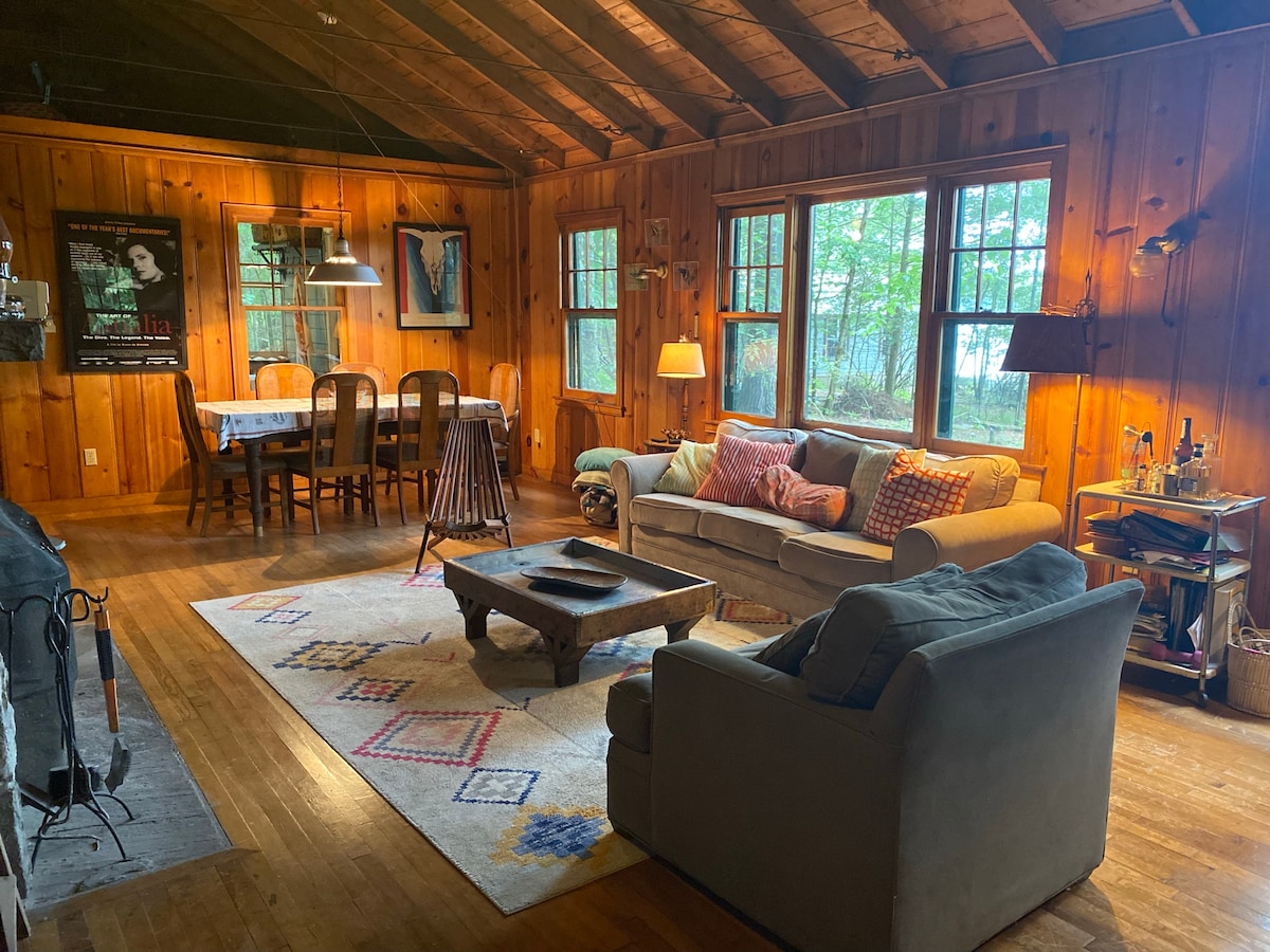 The living area features warm wooden walls and a high-beamed ceiling. A comfortable sofa is positioned with colorful cushions, facing a wooden coffee table on a patterned rug. A dining table for six sits in the background, illuminated by natural light from multiple windows.