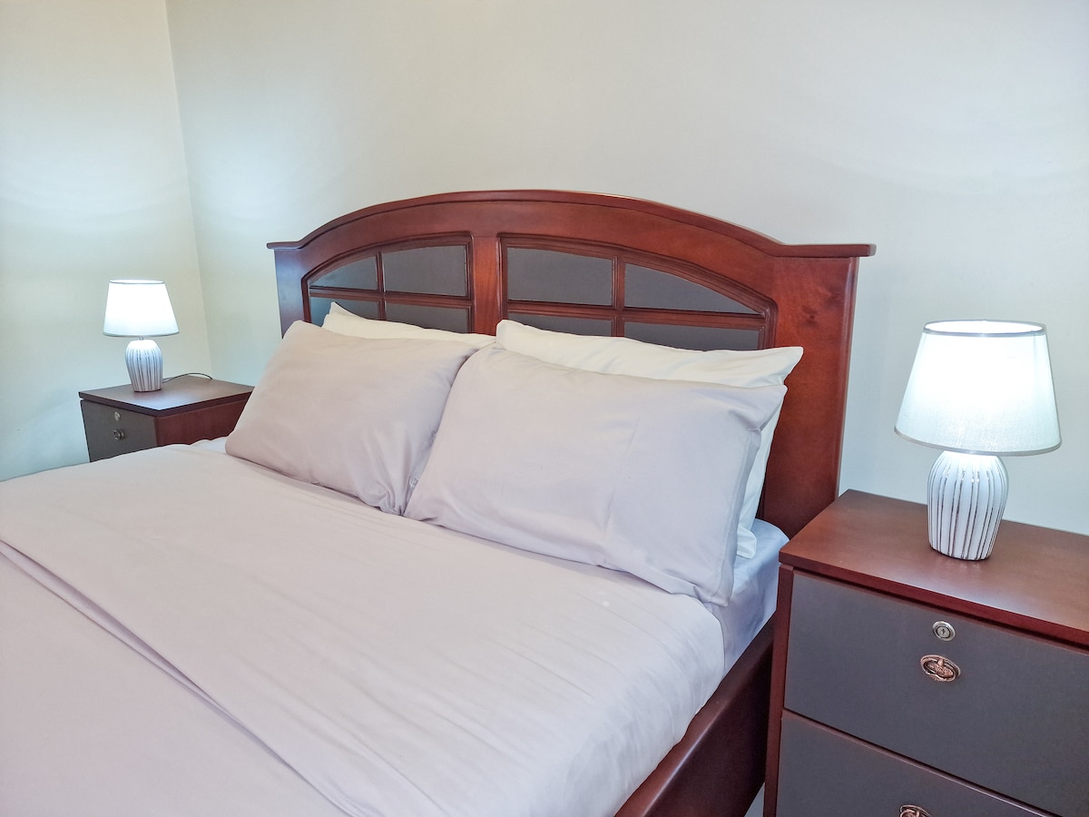 A spacious bed is dressed in neutral-colored linens, flanked by two bedside tables with soft-glowing lamps. The wooden headboard adds a touch of warmth to the inviting sleeping area, which is complemented by a light-colored wall.