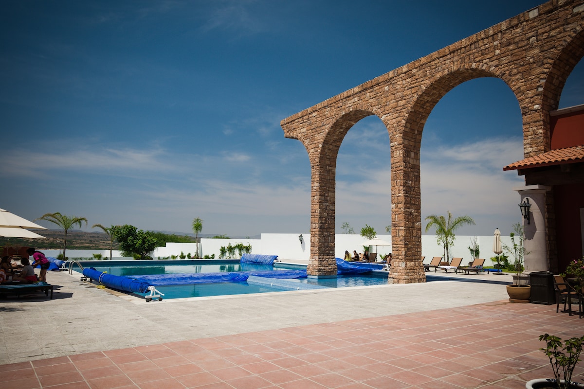 A spacious outdoor area features a large swimming pool, covered partially with a blue tarp. Stone arches frame the scene, and several lounge chairs are arranged alongside the pool. Lush greenery is set against a light-colored wall, enhancing the inviting atmosphere.