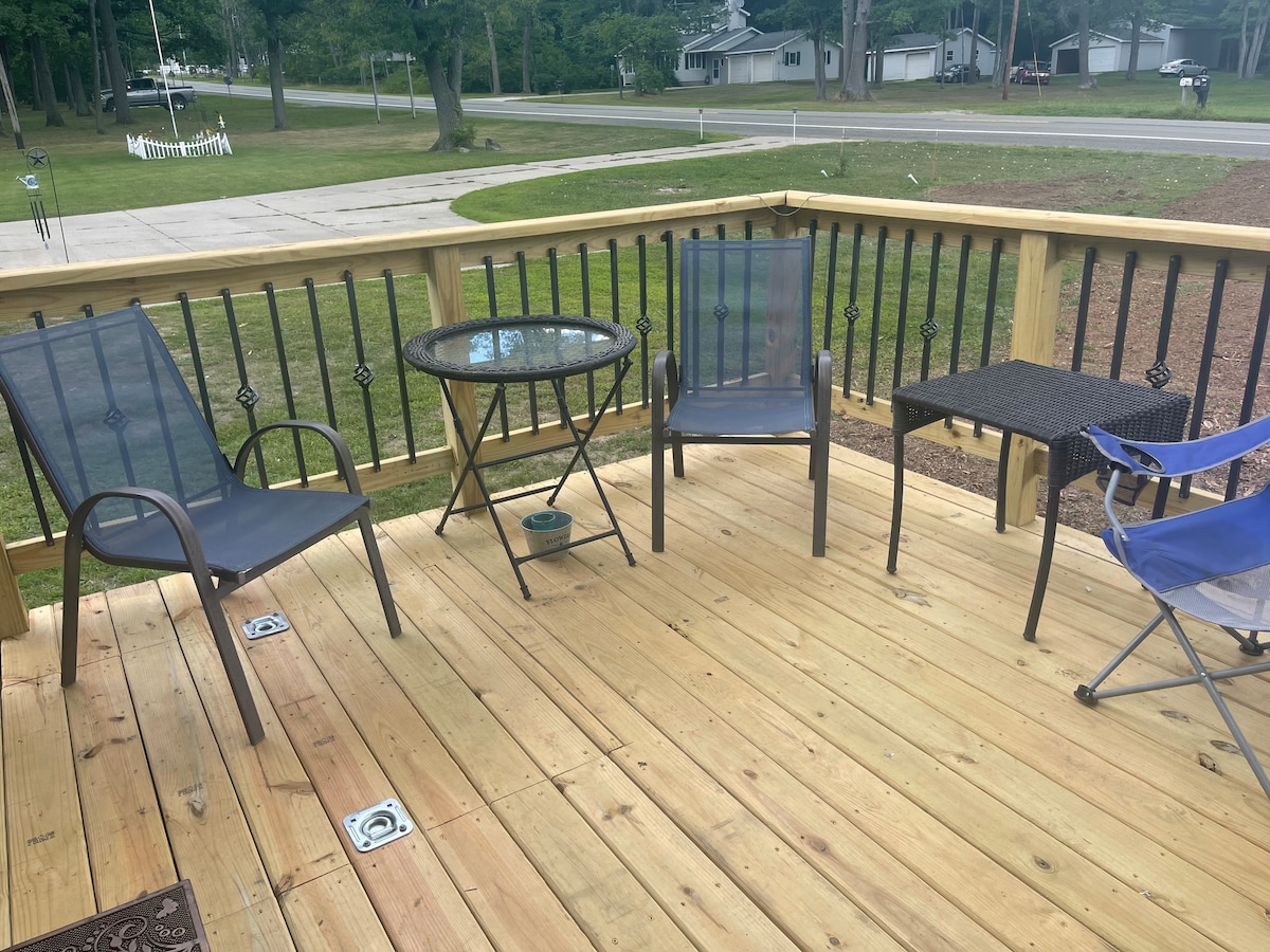 A wooden deck features a circular table surrounded by four chairs. Two of the chairs have cushioned seats, while another is a lightweight folding chair. The deck showcases a spacious layout with decorative railings and is open to a grassy area beyond.