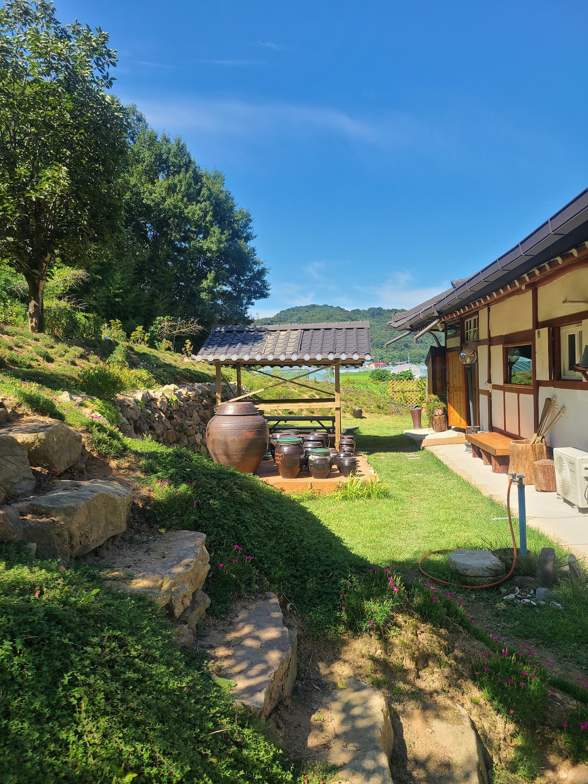 A serene outdoor area is highlighted by a traditional pavilion with a thatched roof, surrounded by greenery. Several earthenware pottery pieces are arranged on a wooden stand. The space offers picturesque views of the landscape and is accessible from the nearby house.