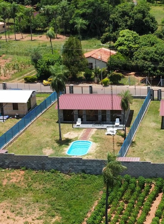 An aerial view of a private garden reveals a compact bungalow with a red metal roof, surrounded by a fenced yard. A small swimming pool is visible, alongside sun loungers on the grassy area. Lush greenery and a gravel path can be seen, enhancing the outdoor space.