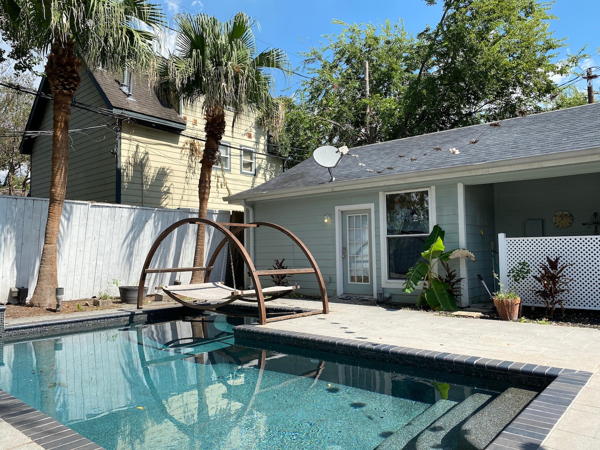 An inviting outdoor space is centered around a sparkling in-ground pool, framed by palm trees and lush greenery. A unique swing set is positioned beside the pool, offering relaxation and leisure. The pale blue exterior of the apartment is visible in the background.