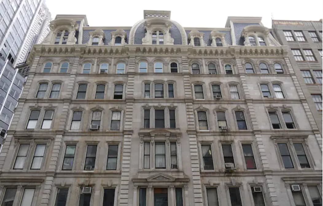 The facade of a historic building is displayed, featuring detailed architectural elements such as ornate cornices and symmetrical window arrangements. Several air conditioning units are visible on the exterior. The structure stands amidst modern buildings, showcasing a blend of old and new architecture.