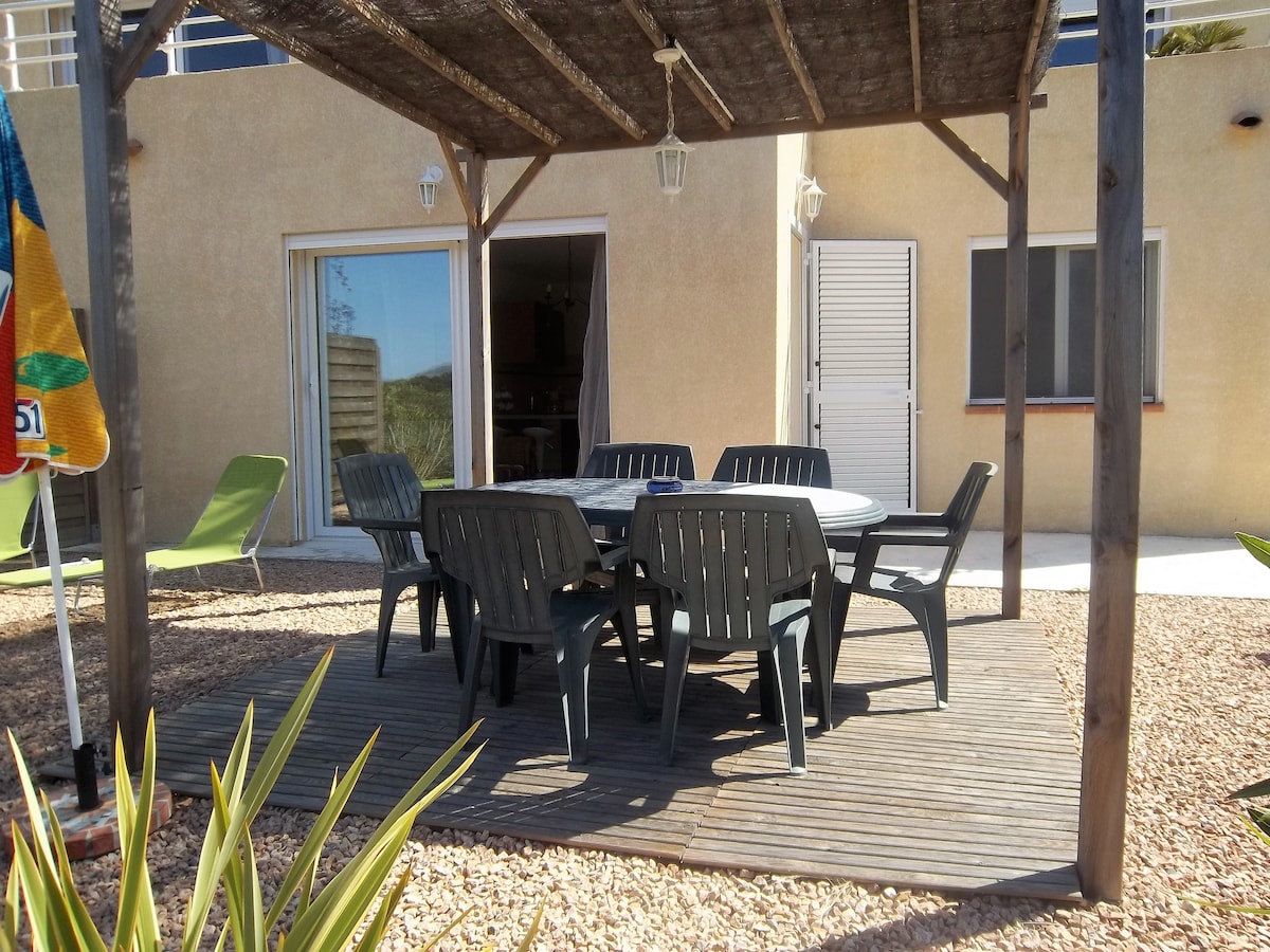 An outdoor terrace features a wooden deck shaded by a rustic pergola. A circular table is surrounded by six chairs, providing ample seating for meals. In the background, light-colored walls and large glass doors are visible, leading into the interior living space.