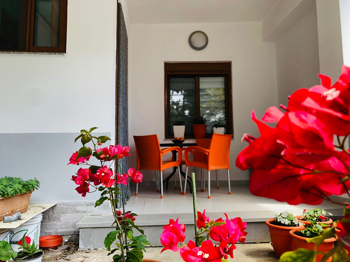 A shaded outdoor patio is presented with an orange table and chairs, inviting relaxation. Surrounding plants and vibrant pink flowers add color, while a subtle gray wall offers a contrasting backdrop. A round light fixture is mounted above the seating area.