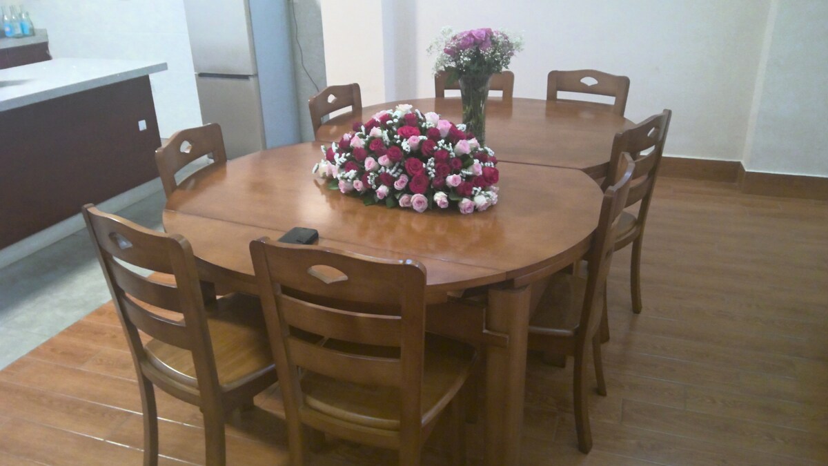 A round wooden dining table is set at the center, surrounded by several matching chairs. A floral arrangement featuring various shades of pink rests at the center, complemented by a simple vase with additional flowers. The wooden flooring adds warmth to the space.