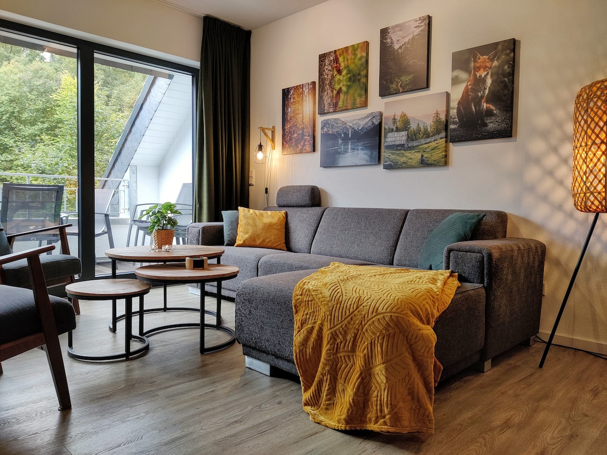 A living area features a spacious grey sofa adorned with a mustard throw. Natural light pours in through large glass doors, enhancing the room's warmth. Two round coffee tables sit in front, and vibrant nature-themed artwork decorates the walls, adding visual interest alongside a floor lamp.