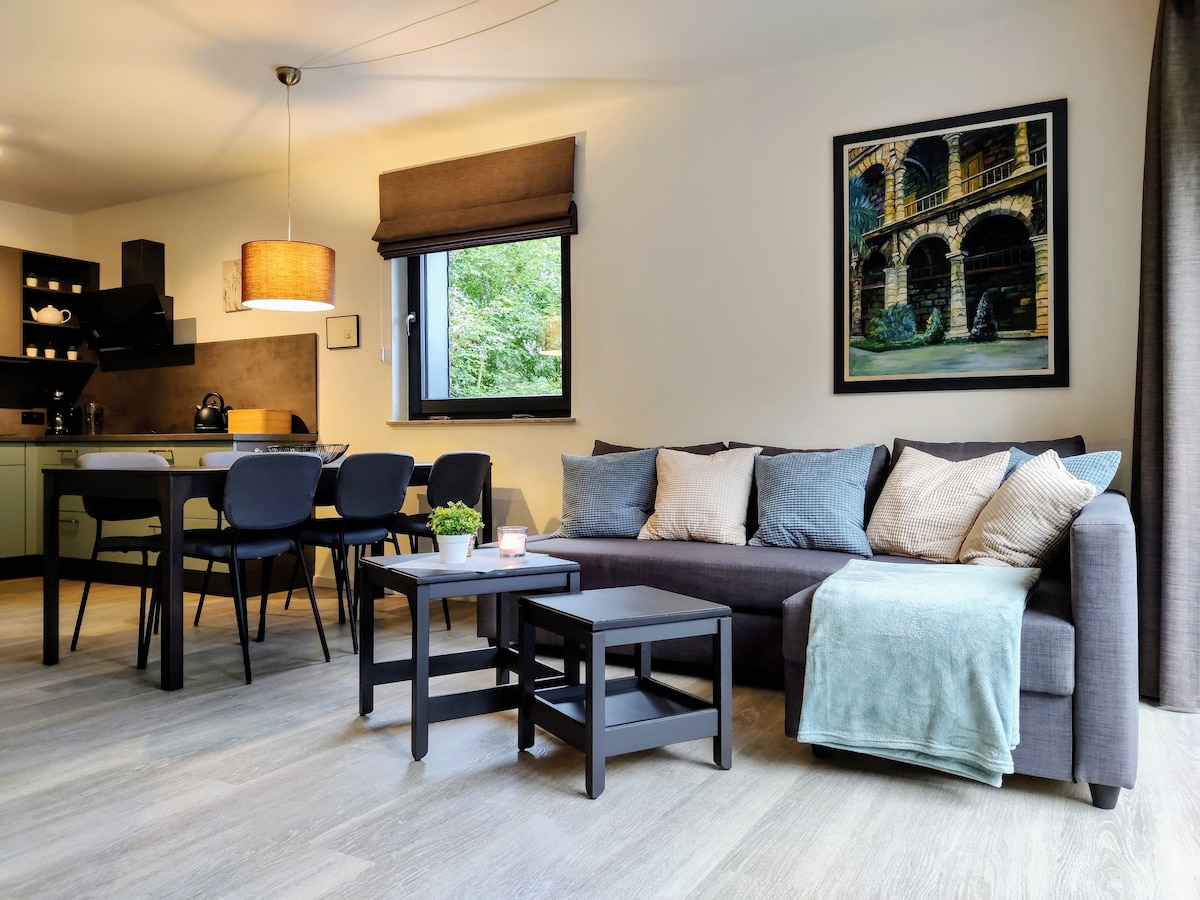 A contemporary living space features a comfortable sectional sofa adorned with decorative cushions. A sleek coffee table sits in front, complemented by a dining area with modern chairs. Natural light is welcomed through a large window, providing views of greenery outside.