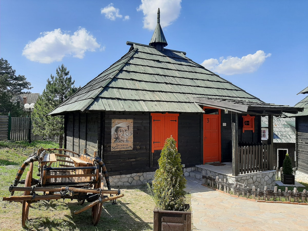 A charming wooden cabin is presented, featuring a distinctive circular design with a peaked roof. The exterior is painted in dark tones, complemented by vibrant orange doors. An inviting pathway leads to the entrance, with decorative wooden features and greenery enhancing the outdoor setting.