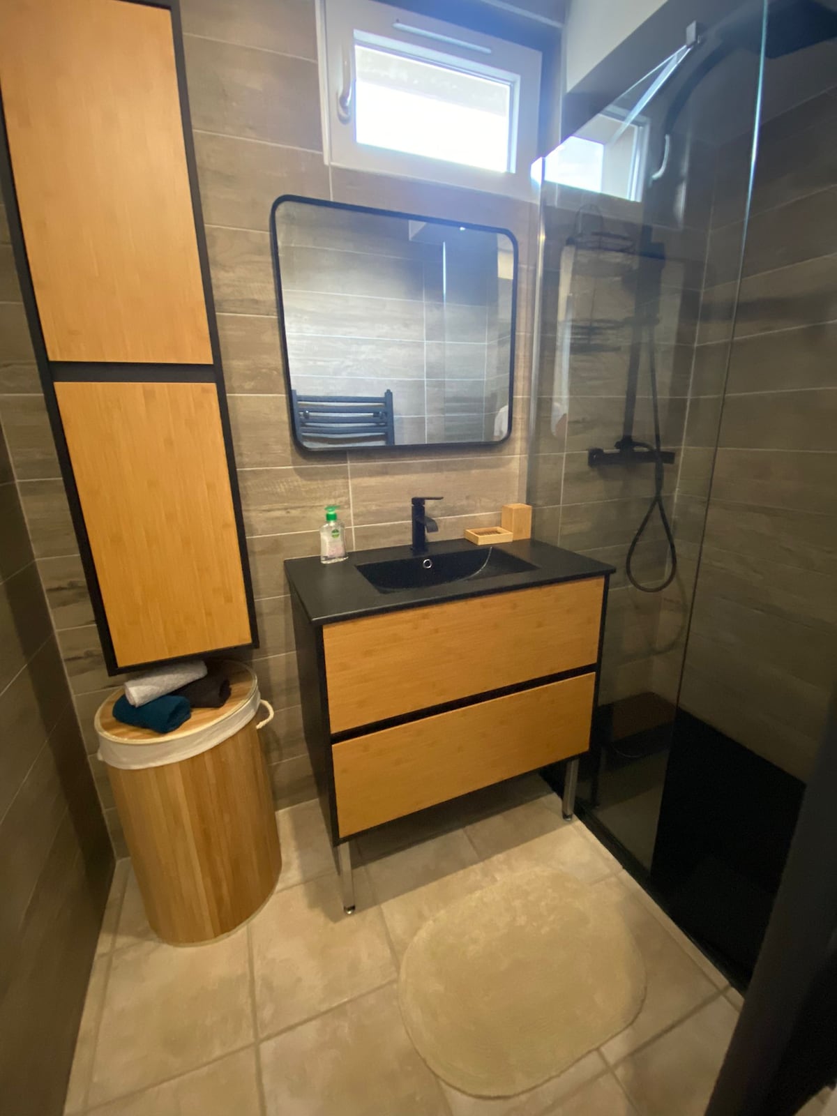A modern bathroom features a clean, minimal design with light beige tiles. A sleek, dark countertop is paired with bamboo cabinetry, including drawers and a tall storage unit. A large mirror is positioned above the sink, and a glass shower enclosure is visible on the right.
