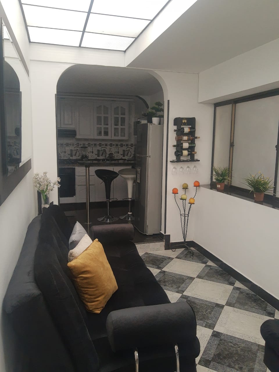 A welcoming living area features a comfortable dark sofa adorned with a yellow pillow. The black and white tiled floor complements the sleek design. A sleek bar with stools is visible in the background, alongside potted plants and decorative wall elements enhancing the space.