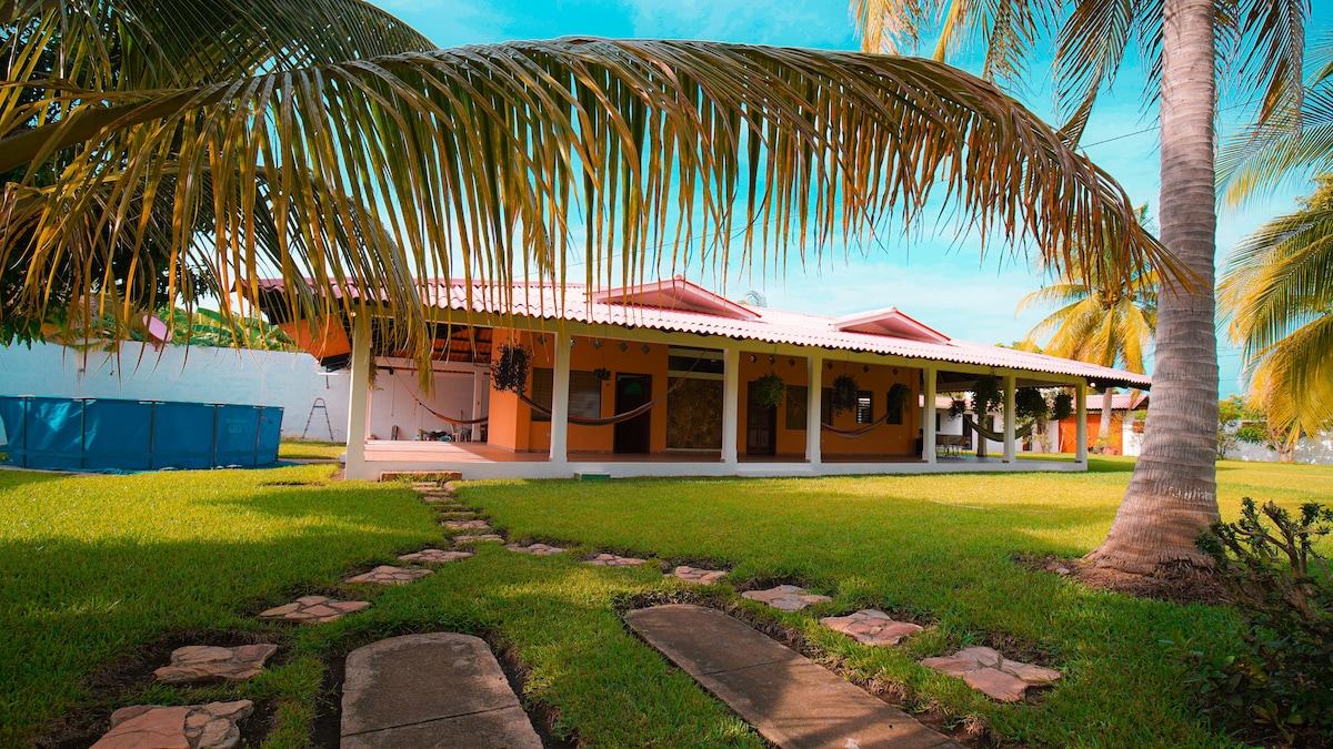 A spacious exterior view of the property is captured, featuring a well-kept lawn bordered by palm trees. Concrete stepping stones lead toward a covered porch, where colorful walls contrast with the serene blue sky above, enhancing the inviting environment.