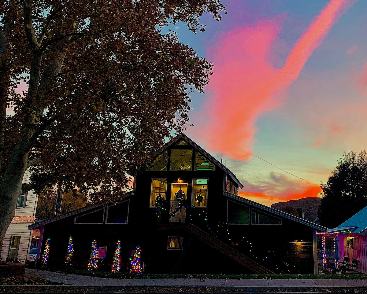 A charming building is illuminated by colorful holiday lights, with vibrant decorations along its exterior. The sky is painted with soft pink and purple hues at sunset. Large windows reflect the changing colors of the sky, suggesting a warm atmosphere.