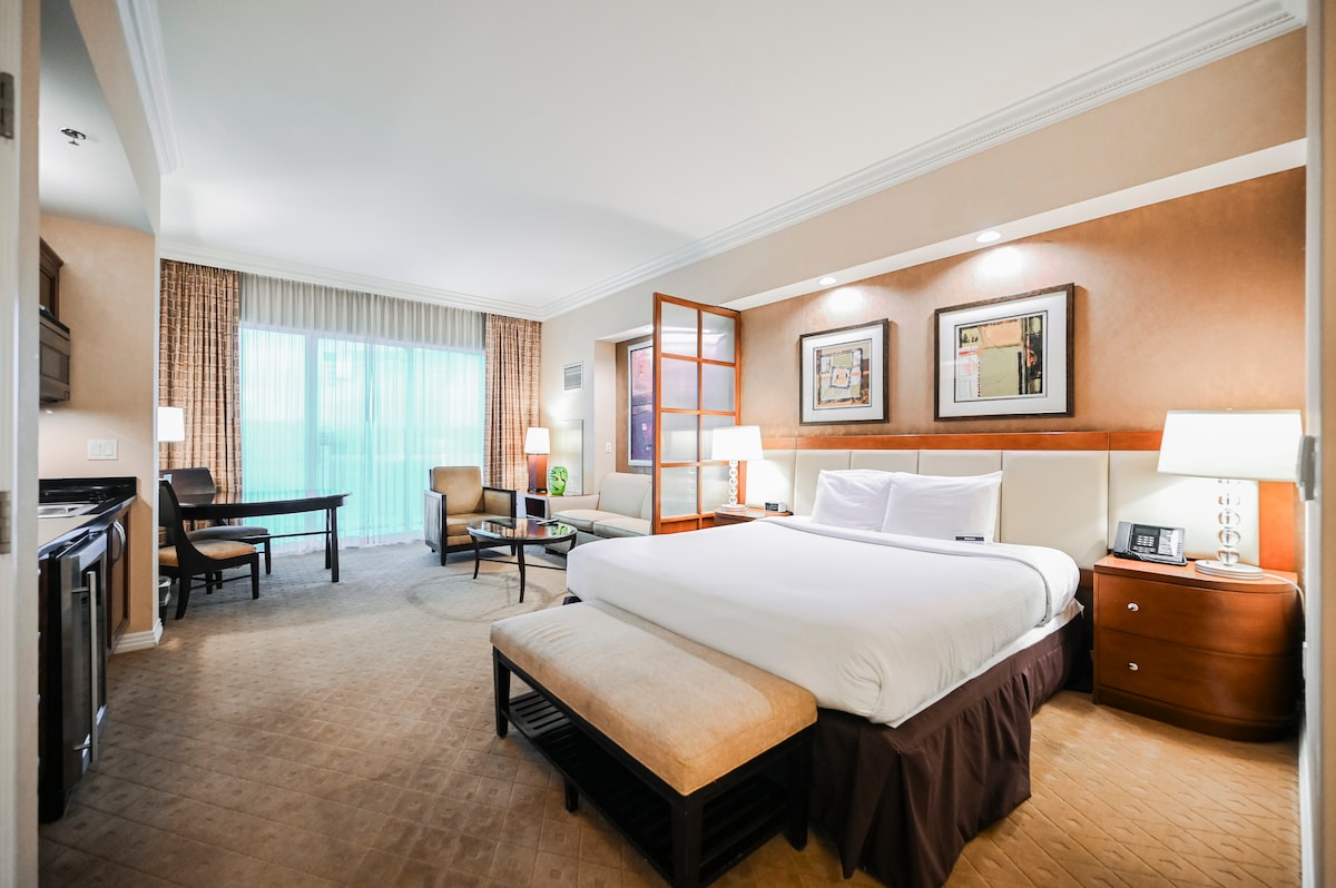 A studio apartment is presented with a plush king-size bed, complemented by a cozy seating area featuring an armchair and sofa. A dining table is positioned near a floor-to-ceiling window draped with sheer curtains, providing ample natural light. A kitchenette is discreetly located along one wall.