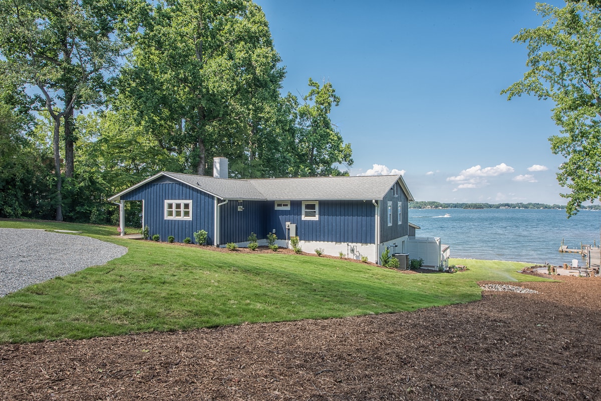 A charming lakefront home is set against a backdrop of lush greenery, with a modern blue exterior contrasting the natural surroundings. A spacious lawn leads to the water's edge, and a driveway is visible, offering easy access.
