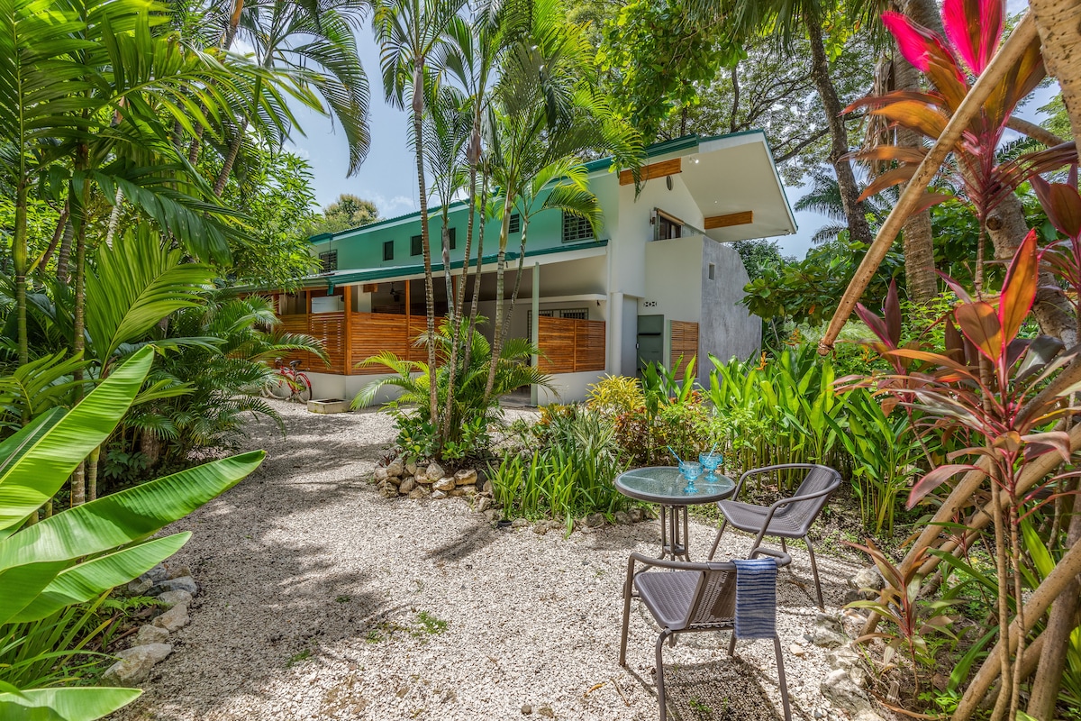 A serene exterior view of the retreat showcases lush tropical foliage surrounding a pathway of white gravel. A small outdoor seating area is positioned on the left, featuring a round table and two chairs. The modern architectural design blends harmoniously with the vibrant garden.
