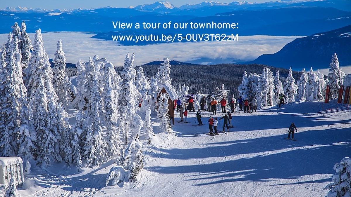 A winter scene unfolds with skiers navigating a snowy slope surrounded by frosted trees. Stunning mountain vistas extend in the background, showcasing a blanket of clouds below, creating a picturesque alpine setting.