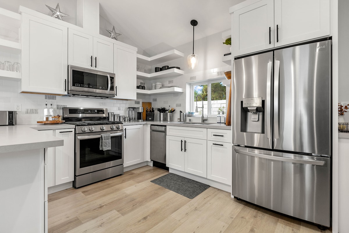 A modern kitchen is presented, featuring stainless steel appliances and a spacious layout. White cabinetry complements the light wood flooring. Open shelves display glassware and cooking essentials, while a large window invites natural light, illuminating the space.
