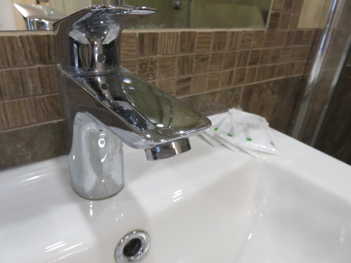 A modern faucet is positioned over a clean white sink, featuring a sleek silver finish. The surrounding walls are adorned with textured brown tiles, complementing the contemporary design. Small packets of toiletries are neatly arranged nearby.