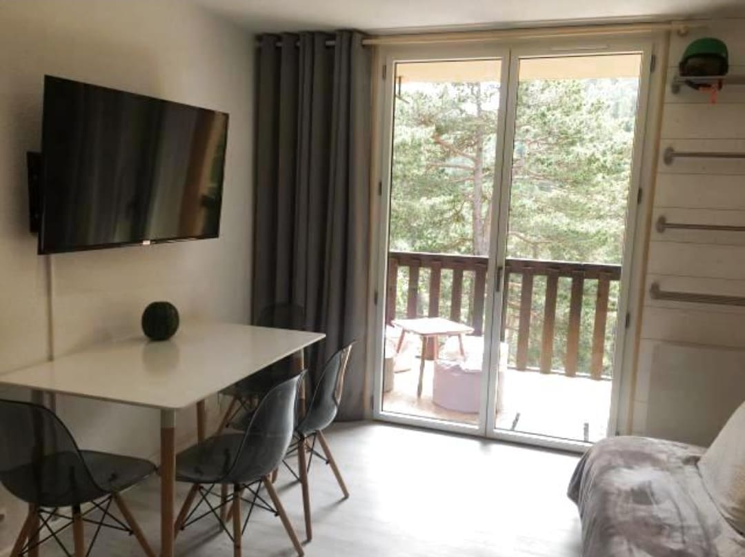 A living area is presented with a large wall-mounted television and a dining table set for four. Sliding glass doors open to a balcony with views of surrounding trees, while soft gray curtains frame the scene, enhancing the natural light.