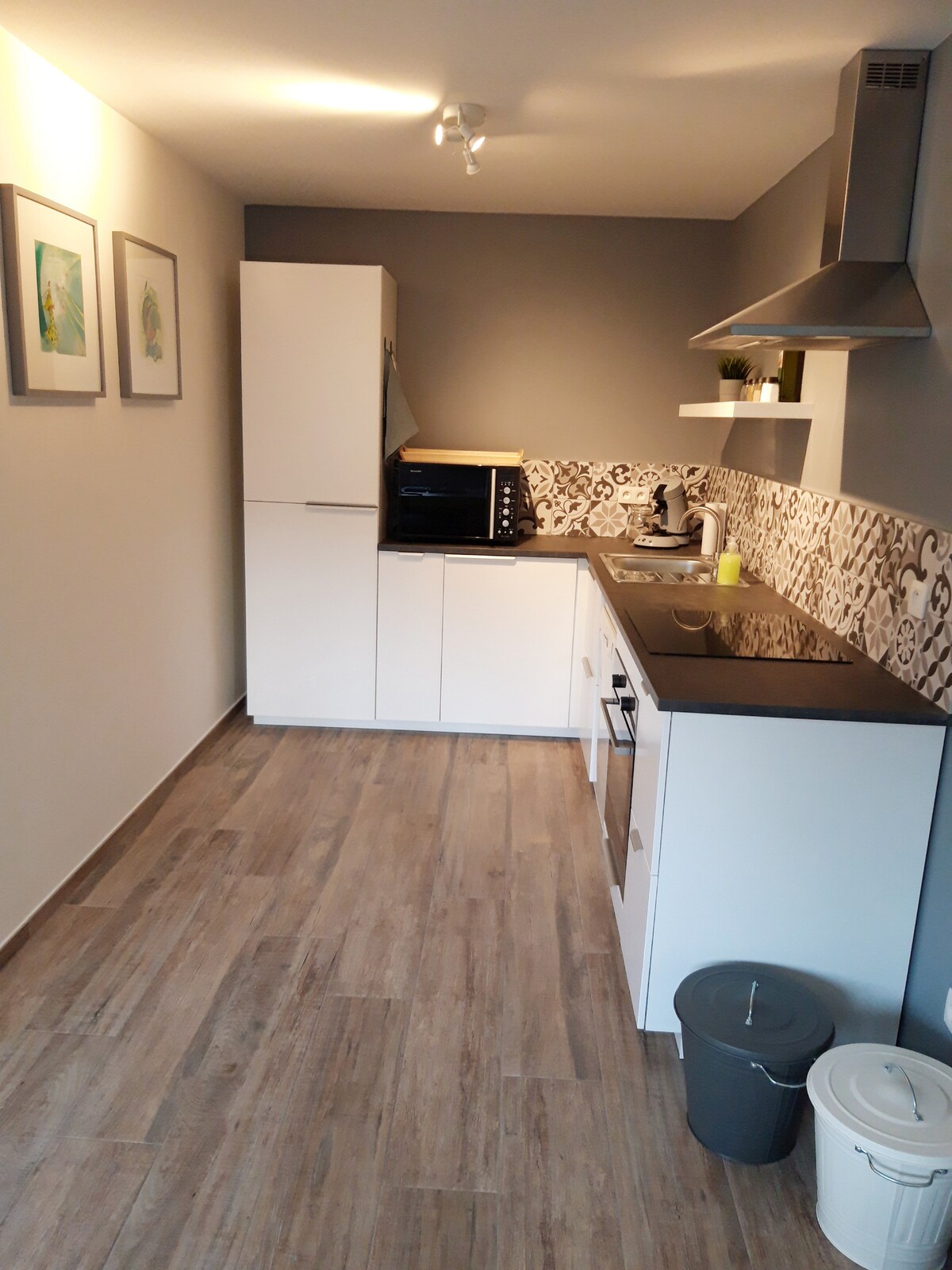 A modern kitchen is presented with sleek white cabinetry and a neutral-toned tile backsplash. Counter space is available for meal preparation, and kitchen appliances, including a microwave and coffee maker, are arranged efficiently. The floor is finished with a light wood effect, contributing to a contemporary feel.