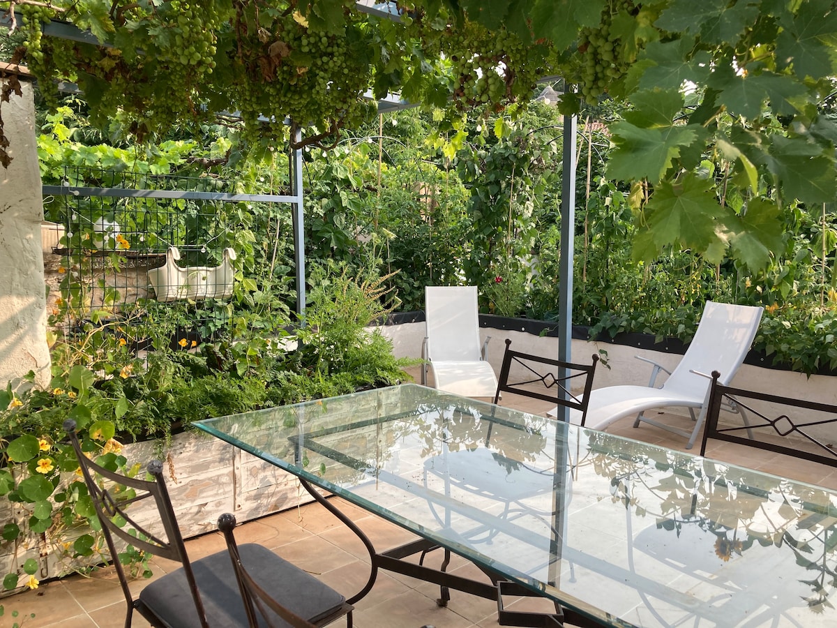 An outdoor terrace is surrounded by lush greenery. A glass dining table is accompanied by four metal chairs, with two lounge chairs positioned nearby. A trellis is seen above, providing shade with vines growing, enhancing the serene environment.