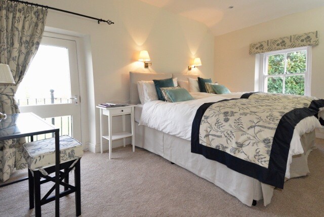 A spacious bedroom is featured, with a large bed adorned with decorative cushions and a patterned bedspread. Soft lighting is provided by wall sconces. A small bedside table and a writing desk with a patterned stool are also visible, enhancing the room's functionality.