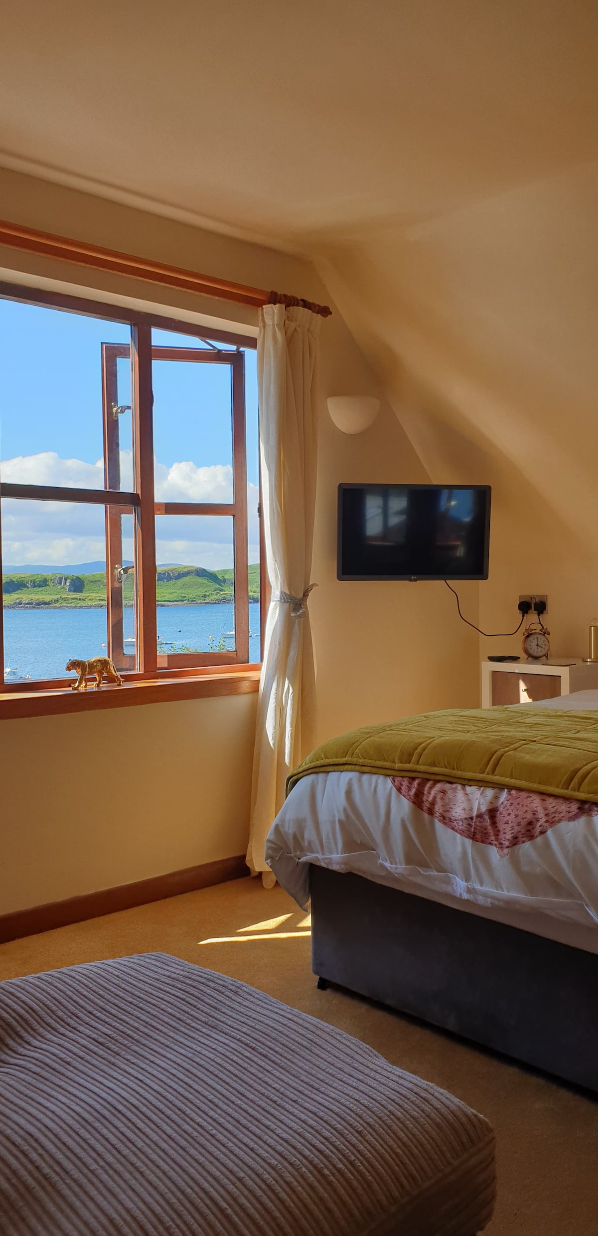 A bright bedroom features a spacious bed with a warm throw and a large window offering scenic views of the water and surrounding landscape. A wall-mounted television is visible, and a small chair is positioned nearby. Natural light fills the room, enhancing its airy feel.