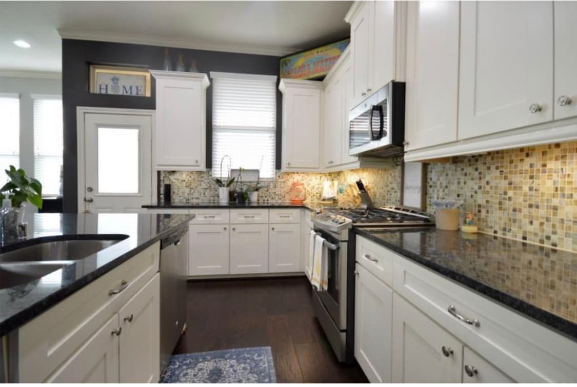 A modern kitchen features white cabinetry and a dark granite countertop. A mosaic tile backsplash adds texture and color. Natural light illuminates the space through large windows, while stainless steel appliances are integrated for functionality. A door leads to the exterior.