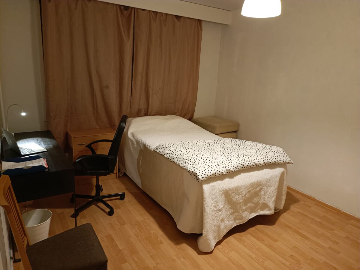 A minimalist bedroom is presented featuring a spacious bed covered with a white duvet and a polka dot blanket. A wooden chair and desk are included, along with a small spot lamp providing soft light. A curtain drapes partially over the window, contributing to a serene environment.