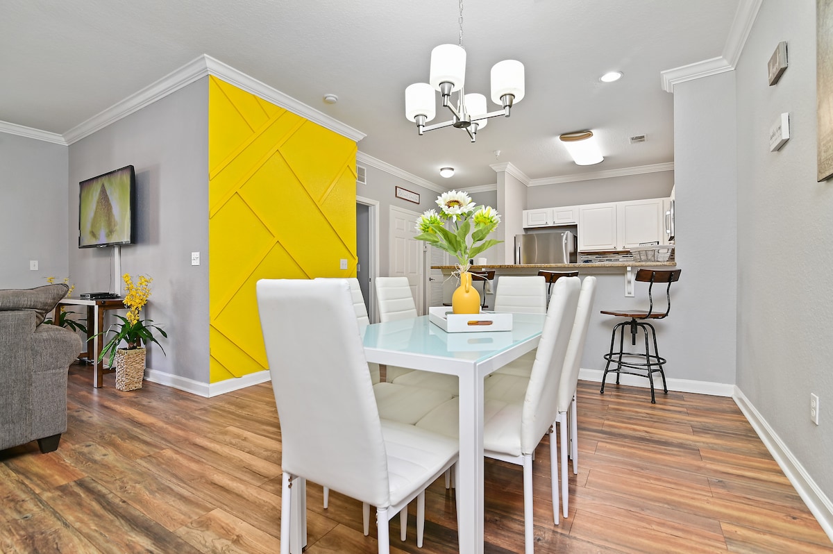 A dining area is shown with a modern white table surrounded by six white chairs. A floral centerpiece adds a touch of color, while a bright yellow accent wall and a mounted television enhance the lively atmosphere. The open layout connects to a living space and kitchen.
