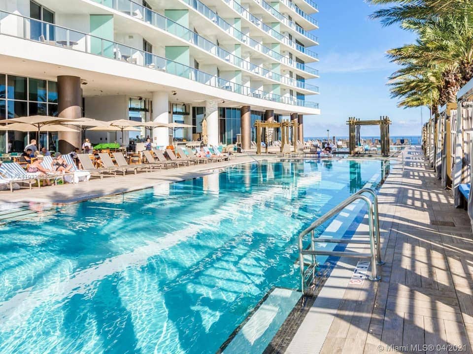 A spacious outdoor pool area features clear blue water complemented by comfortable lounge chairs and private cabanas. Palm trees line the poolside, adding a tropical touch, while modern architecture creates an inviting atmosphere. The ocean view is visible in the background, enhancing the resort experience.