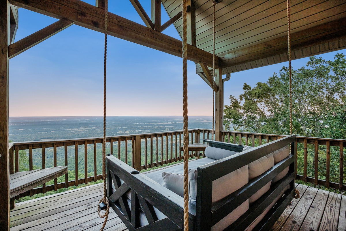 A hanging daybed swing is positioned on a spacious wooden deck, offering expansive views of the valley below. The deck is surrounded by wooden railings, creating a serene space to relax and enjoy the natural surroundings.
